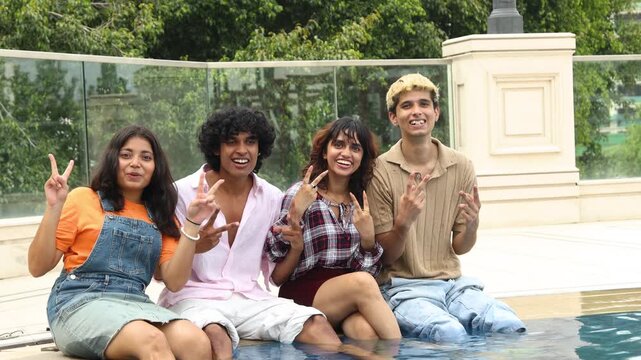 Diverse young indian asian Gen Z friends sitting together with their legs in swimming pool enjoying happy summer day outdoors. Travel and Holidays..Youth Culture.
