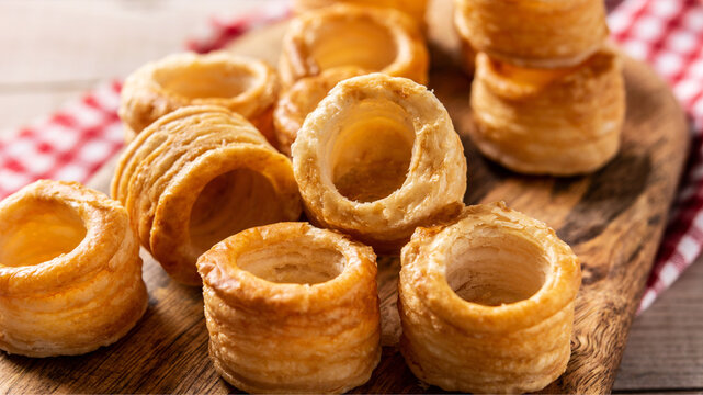 Golden flaky puff pastry shells arranged on a rustic wooden board, showcasing delicate layers and a hollow center, perfect for savory fillings and elegant appetizers.