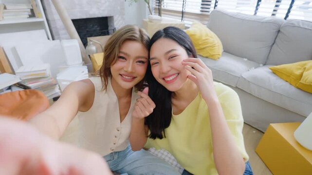 Two young Asian women smiling and happy while taking a selfie in their living room at home, looking at the camera