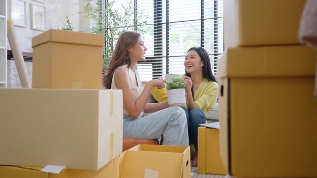 Happy two Asian women friend picking up things out to a paper box while preparing to move in of the new home. Relocation concept