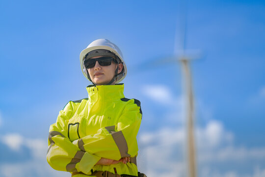Portrait of a woman in wind power standing quietly in her yellow jacket with hands clasped low and a single blurred turbine tower rising at the right of frame, green electricity at source
