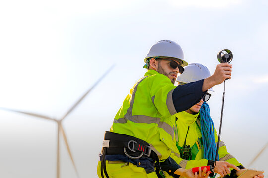 Rear view of a technician holding the anemometer aloft with the cable hanging in a long curve, a turbine tower rising directly behind his raised right arm as another quiet workday unfolds