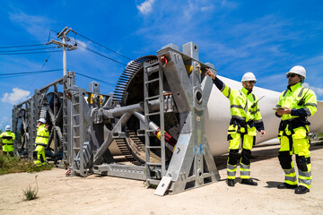 Fototapeta premium An engineer points up at the pitch bearing teeth of a wind turbine hub as his colleague makes notes on a tablet beside him at a green energy construction site in tropical daylight.