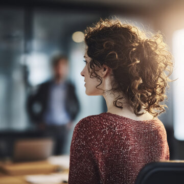 Rear view of a businesswoman listening during an office meeting. A colleague presents ideas in the blurred background.