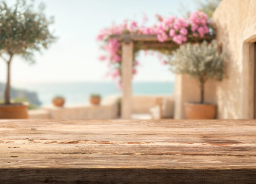 Wooden tabletop frames blank display area against blurred coastal terrace. Sunlit rustic boards stay central, while out of focus sea, potted greenery, and flowers support airy summer lifestyle