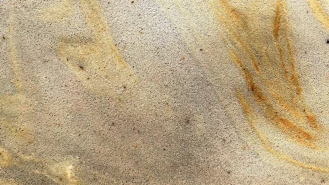 Thin foamy water film drifts across sandy surface with orange mineral streaks