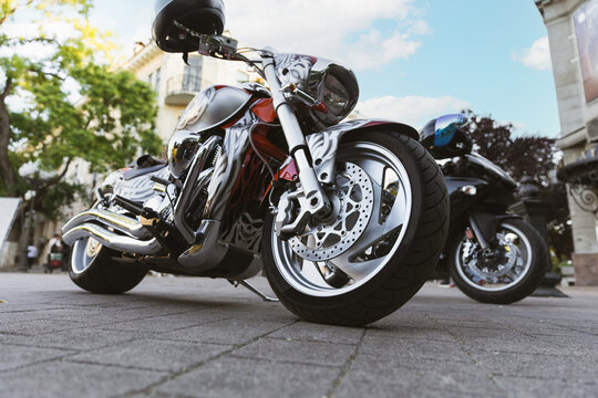 Custom motorcycle rests on city pavement in urban showcase. Low angle view highlights chrome wheel, front brake, and bold paint while another bike and buildings frame parked machine.