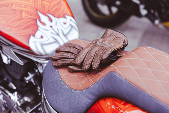 Gloved hands rest on motorcycle seat. Custom stitched upholstery and brown riding gloves create stylish closeup of parked bike, with polished bodywork adding bold urban feel beside saddle stitching.