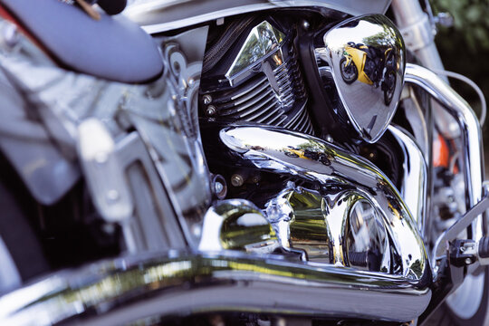 Chrome motorcycle engine fills frame with polished metal reflections. Motorcycle detailing stands out in bright daylight, while mirrored surfaces show nearby bikes and trees on stationary machine.