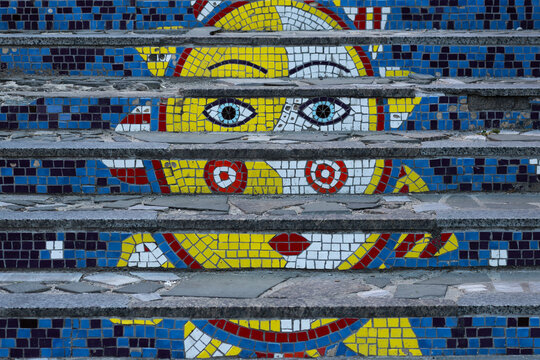 Decorative mosaic steps show stylized eyes, lips, and abstract shapes. Weathered stone stairs are lined with vivid blue, yellow, red, and white tiles in vibrant artistic urban setting.