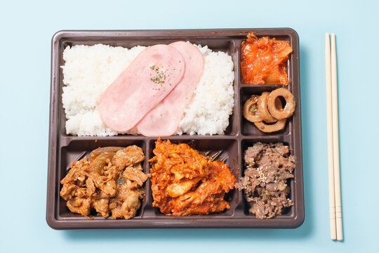 Korean lunch box with rice, meat, and kimchi in plastic packaging on a blue background close-up, top view.