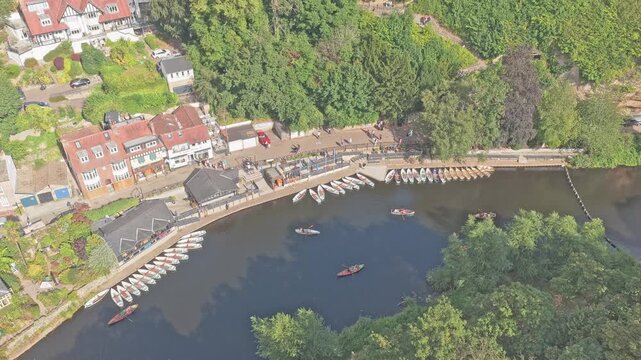 Rowing boats and moored skiffs line the River Nidd waterfront beside riverside buildings and woodland in Knaresborough, North Yorkshire, England, United Kingdom, drone forward flyover with tilt down.