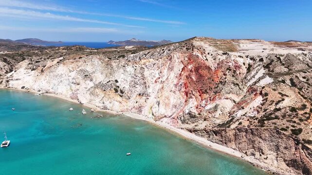 Firiplaka Beach stretches below layered red, ochre and white volcanic cliffs, with pale sand, clear turquoise water and rocky shoreline on Milos, Cyclades, Greece, drone slide