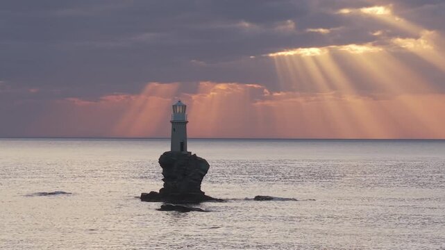Drone orbit around the historic Andros Lighthouse at sunrise, capturing golden sun rays streaming through the sky and creating a shimmering path of light across the Aegean Sea.