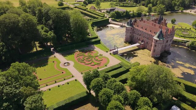 An aerial reveal of Egeskov Castle, highlighting its distinctive architecture and surrounding ornamental gardens, lawns, trees, and landscaped estate.
