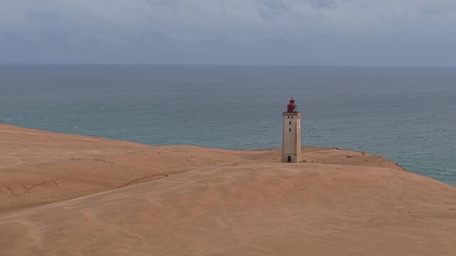 Aerial footage circling Rubjerg Knude Lighthouse on a cloudy day, showcasing expansive sand dunes, the rugged North Sea coastline, and the dramatic landscape of Northern Jutland, Denmark.