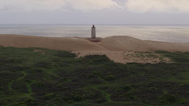 Aerial footage revealing lush green grass that transitions into the sandy dunes surrounding Rubjerg Knude Lighthouse along Denmark's dramatic North Sea coastline.