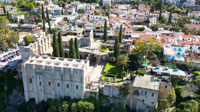 Aerial drone footage of Bellapais Abbey, a historic Gothic monastery in Bellapais village near Kyrenia, Cyprus, surrounded by buildings, trees and the Mediterranean landscape.