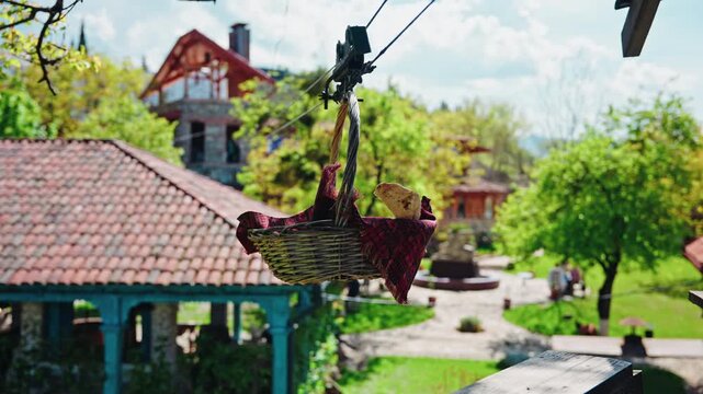 A wicker basket lined with a red patterned cloth hangs from a rope line above the tiled roofs and garden of a Georgian winery.