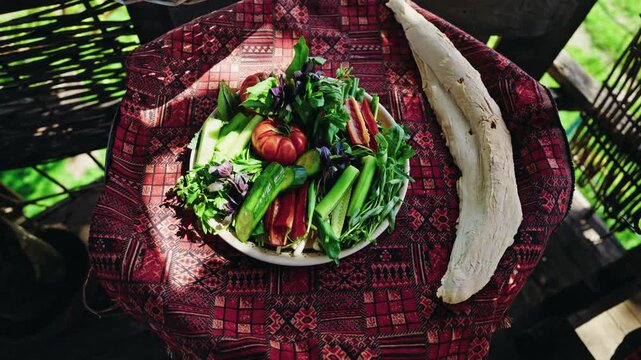 Overhead view of a plate of fresh vegetables and a sheet of lavash bread laid on a red patterned carpet over a wine barrel.