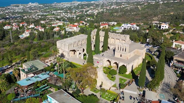 Drone aerial footage showing the Bellapais Abbey complex from above, with historic stone buildings, courtyards, surrounding vegetation and the nearby village visible in the frame.