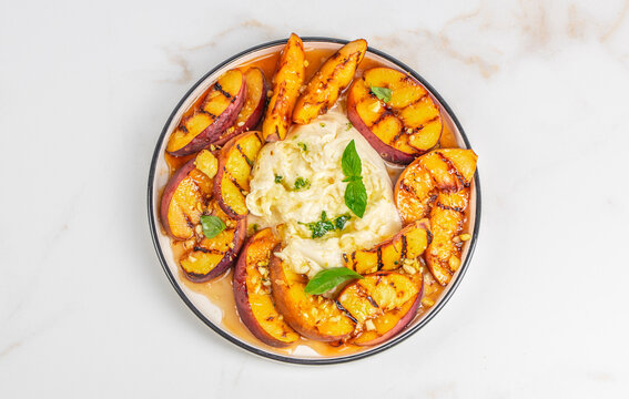 Grilled peaches arranged on a plate with creamy ricotta cheese and fresh basil leaves, set against a light marble background