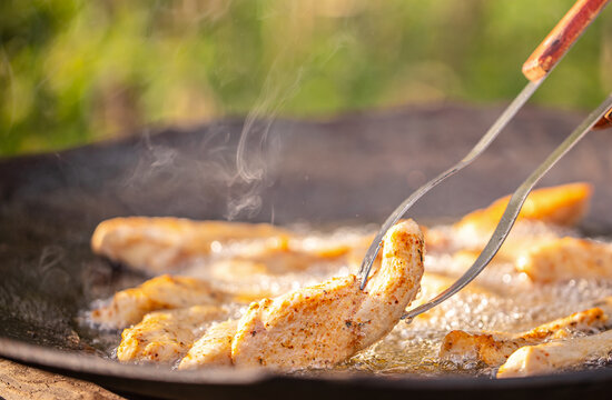 Grilled chicken fillets being cooked in a cast iron skillet over an open flame, with steam rising and a blurred green background of outdoor foliage