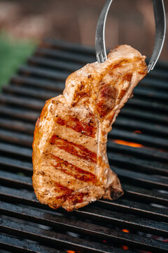 Grilled pork chop being lifted with tongs from a barbecue grill, showcasing charred grill marks and juicy texture against a blurred outdoor background