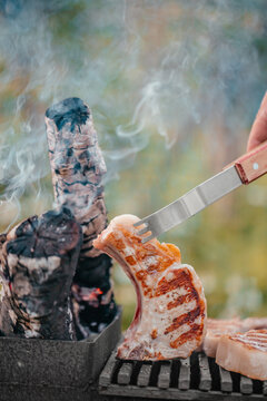 Grilled meat being lifted with a fork from a barbecue grill with smoke rising from charred wood logs in the background during an outdoor cooking session