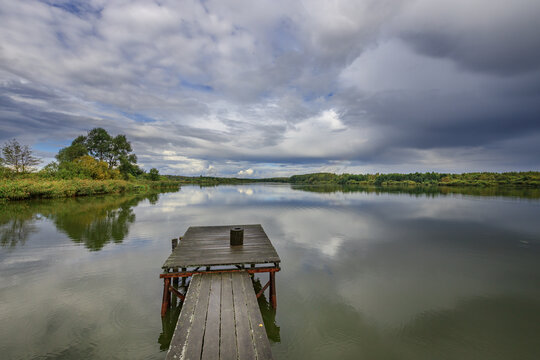 Weathered pier leads into serene lake, framed by trees under moody clouds