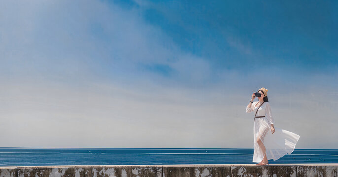 The a woman in a flowing white dress photographs the scenic coastal background against a vast blue sky and ocean view.