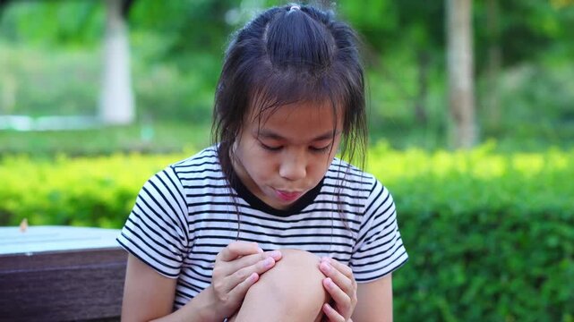 Sad Asian girl sitting and touching injured bleeding knee with painful expression outdoors