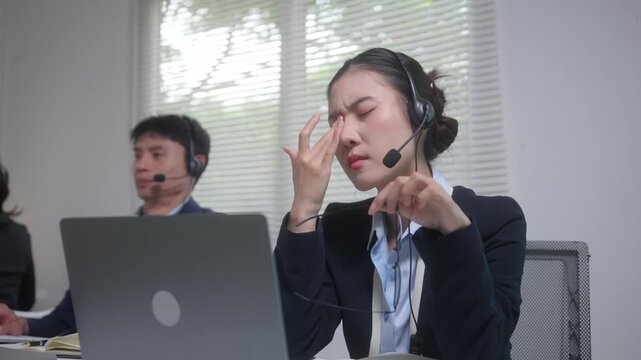 Stressed call center operator feeling eyestrain and fatigue from overworking with a laptop and wearing a headset in the office