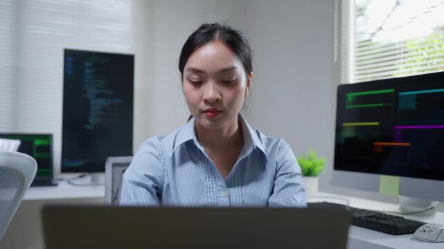 Asian female developer writing code and programming on a laptop while smiling at the camera in a modern tech office