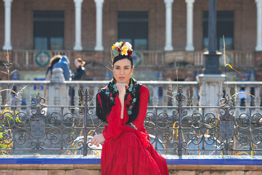 A beautiful flamenco dancer sitting on a tiled bench in a square in Seville, Spain. She is wearing a red ruffled dress and has a flower in her hair. Flamenco, a UNESCO Intangible Cultural Heritage.