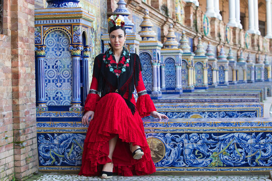 A beautiful flamenco dancer sitting on a tiled bench in a square in Seville, Spain. She is wearing a red ruffled dress and has a flower in her hair. Flamenco, a UNESCO Intangible Cultural Heritage.