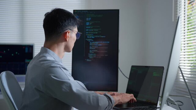 Asian programmer coding on a computer writing software and programming a new script in a modern office with multiple displays