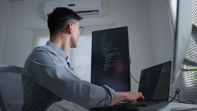 Asian male programmer coding and developing new software at his desk in a modern office with multiple monitors