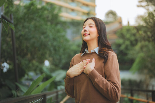 Peaceful young woman standing outdoors with eyes closed and hands over heart, practicing gratitude and mindfulness in nature, expressing inner peace.