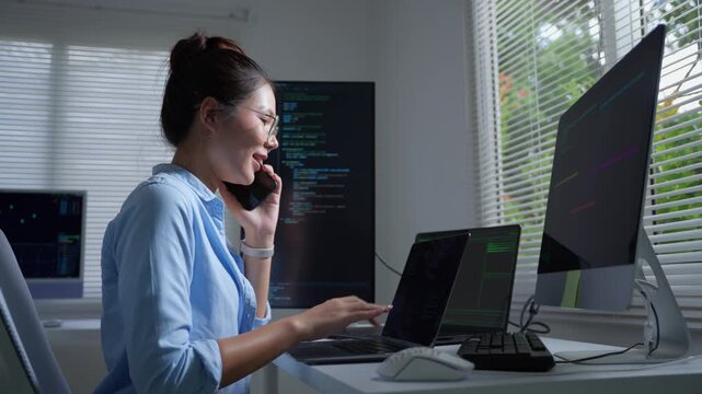 Asian software developer working on computer and talking on phone while coding in a modern office