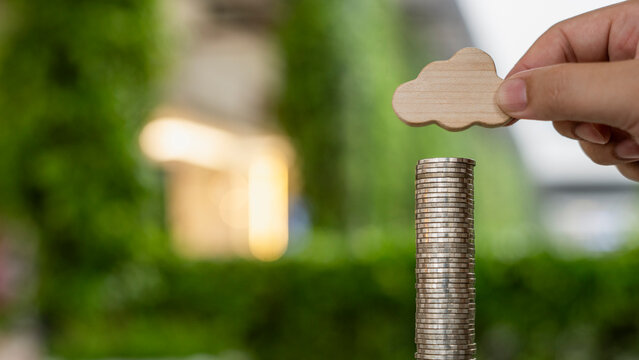 Cloud computing investment cost and digital technology funding concept with hand holding wooden cloud icon above stacked money coins in modern office background.