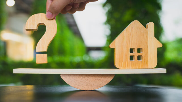 Real estate property decision concept with wooden house model and question mark balancing on seesaw scale, representing housing choices, mortgage loan dilemma, and home investment planning.