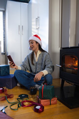 Obraz premium Adult female sitting on floor near stove, wearing Santa hat, holding smartphone, wrapping gifts