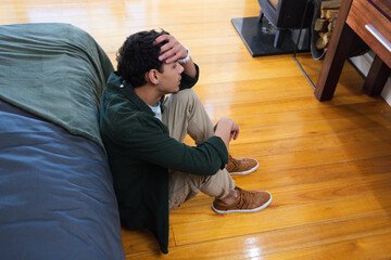 Obraz premium African American man sitting on wood floor beside bed, holding head, looking down by stove
