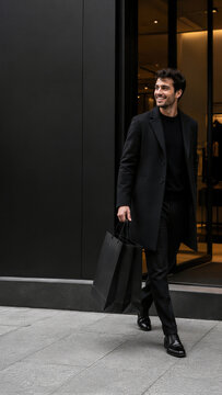 Smiling man in black coat walking with shopping bags near modern store. Elegant male shopper leaving boutique on city street. Black friday sale and shopping concept.