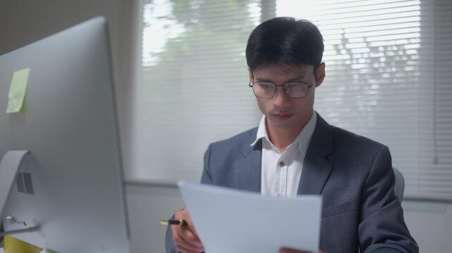 Young professional employee in glasses analyzing paperwork and data on the screen. A dedicated worker comparing financial reports