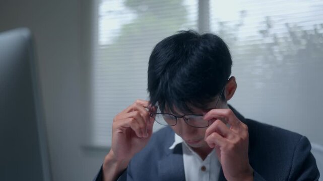 Tired asian businessman feeling eye strain and headache from overworking on computer in office at night