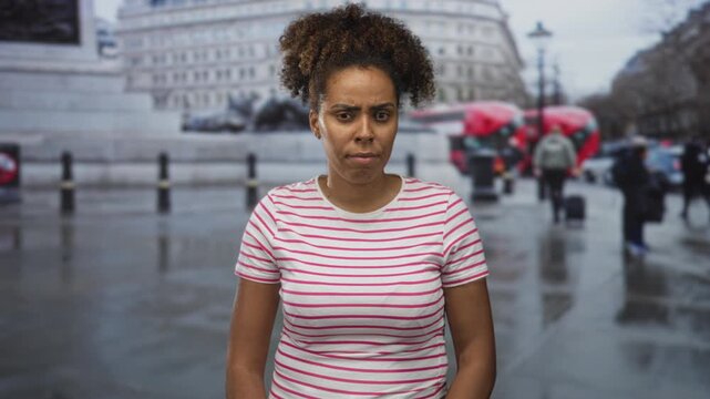 Woman with bare face frowning while standing on a rainy street near a red bus and stone building; travel frustration.