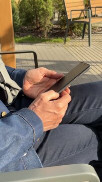 male user holding smartphone and texting message on city square bench. Mature caucasian man types text document on mobile phone display outside. Contemporary portable electronics enable