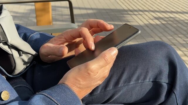 Browsing fast digital media content on smartphone touch screen outdoors. Mature person uses mobile internet app to scroll social network feed on bench. Doomscrolling concept explores